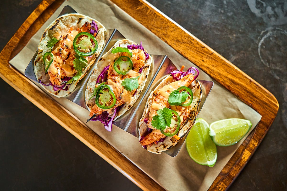three spicy chicken tacos with fresh jalapeno and lime slices on the side on a wooden tray