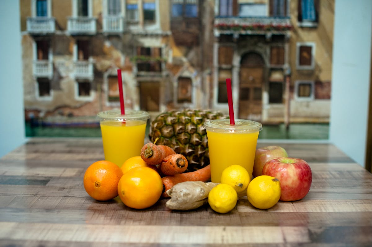 two orange juices sitting on a wooden table with other fruits 
