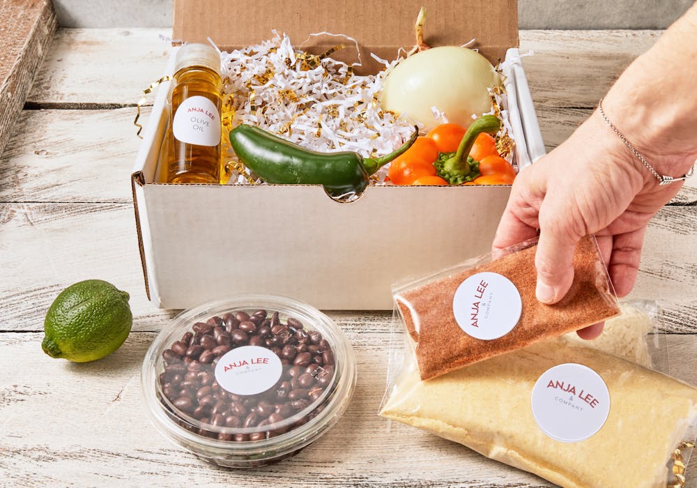 a hand grabbing a bag of spices surrounded by a container of beans, vegetables, and olive oil all packaged in a white box