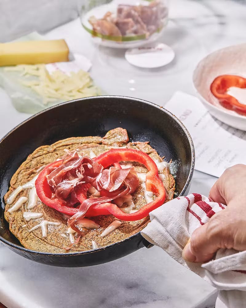 a crepe in a black pan topped with cheese, proscuitto, and a sliced red pepper