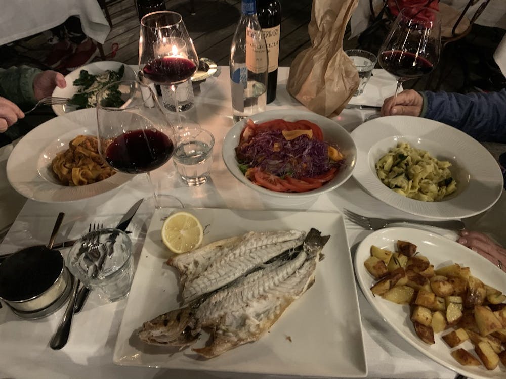 plates of fish, pasta, and vegetables on a white tablecloth table with glasses of red wine