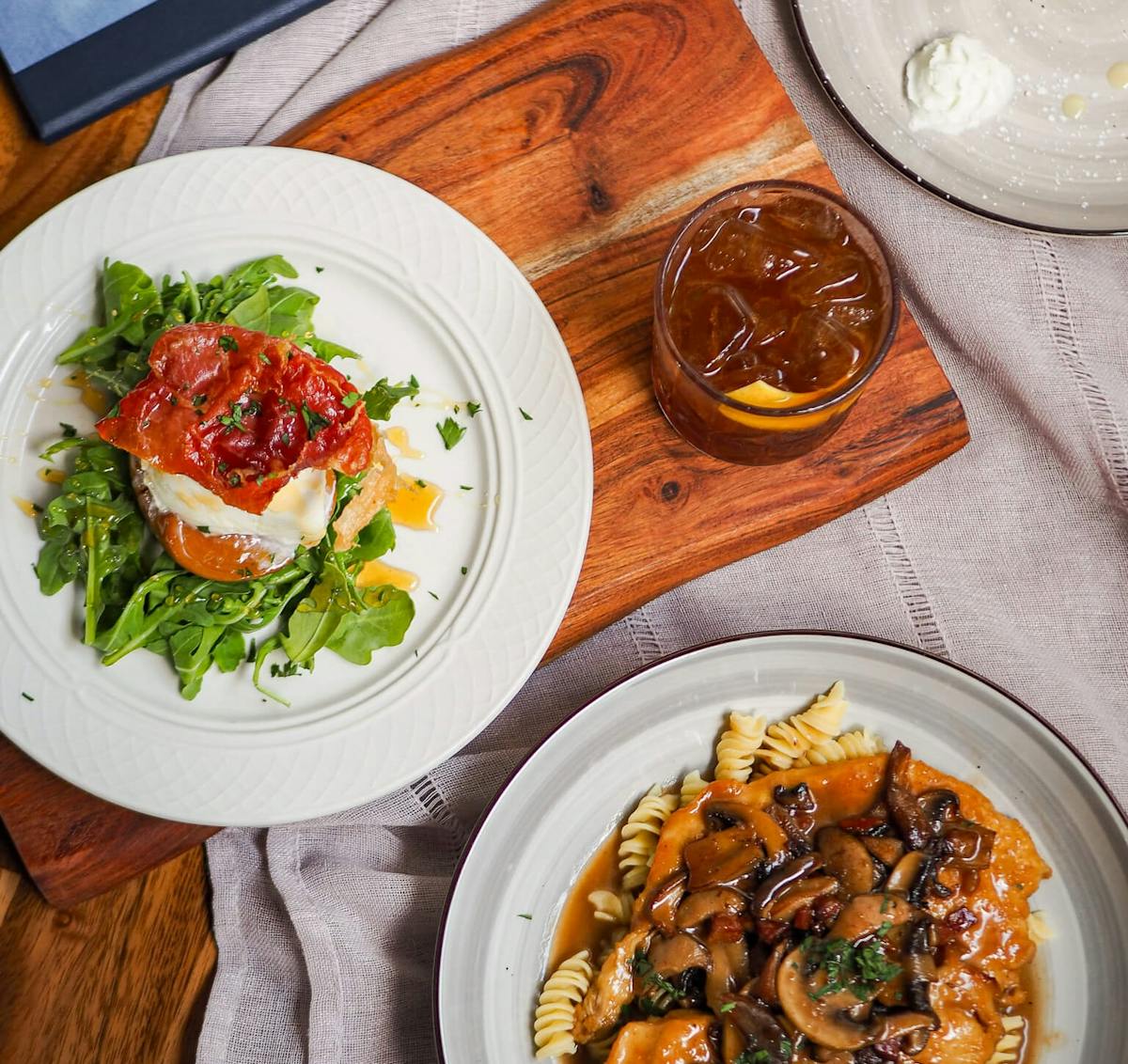 Overhead view of arugula salad with prosciutto, mushroom chicken pasta, tiramisu dessert, and a cocktail at Vuolo restaurant