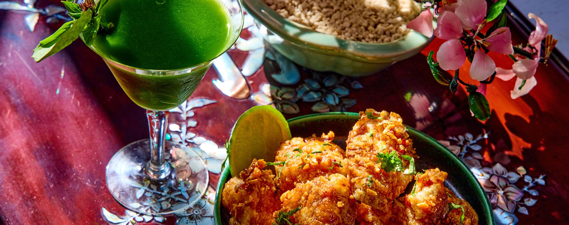 A green cocktail next to a bowl of fried food, set on a floral-inlaid tray for a Grove pop-up dinner.