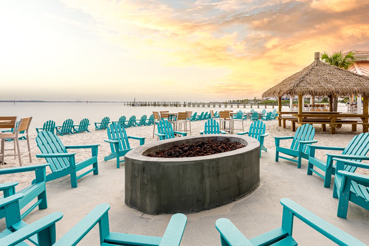 a fire pit and chairs on the beach during sunset